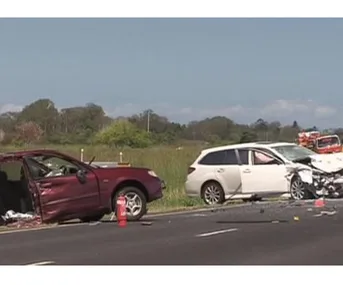 Traffic accident scene with two severely damaged cars on a highway, emergency vehicle visible in the background.