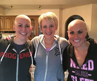 Three sisters, two with shaved heads, smiling in a kitchen, wearing casual hoodies.