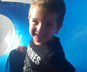 A young boy smiling, wearing a black Ramones t-shirt, standing near a blue play structure.