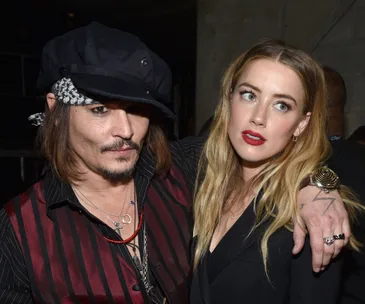 Two individuals posing together at an event; one wearing a hat, the other with long blonde hair.