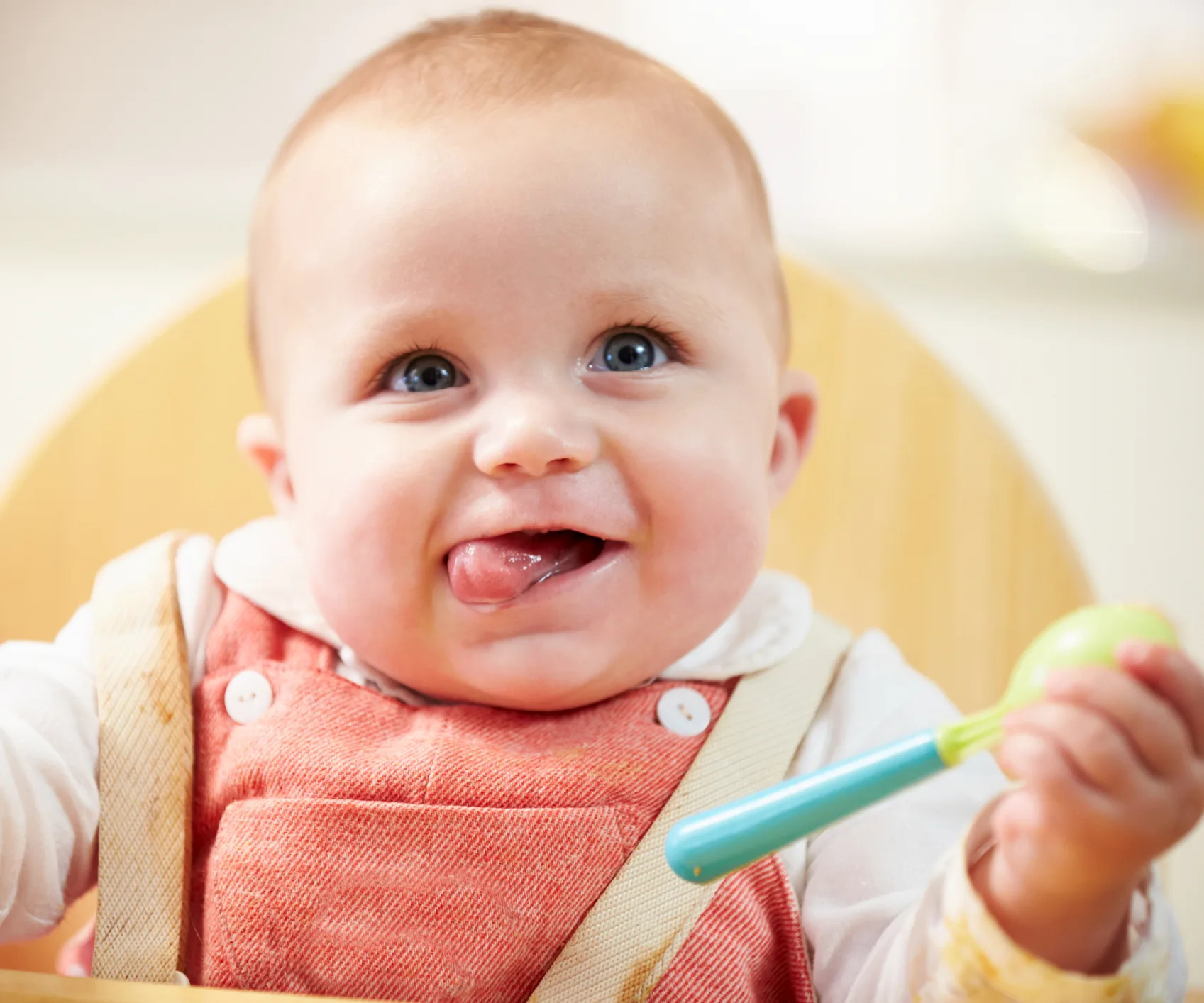 A smiling baby wearing a pink bib sits in a highchair holding a green and blue spoon.