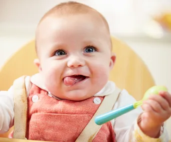 A smiling baby wearing a pink bib sits in a highchair holding a green and blue spoon.