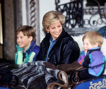 A woman smiles with two young boys sitting on a sled, all bundled in colorful winter jackets.