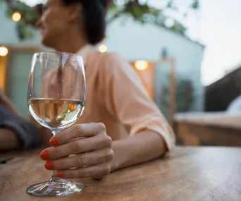 A woman with red nail polish holds a glass of white wine at a wooden table outdoors.