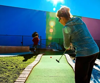 Person playing mini-golf under a bright sun, with a child in the background near a colorful building.