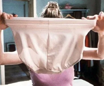 A woman holding up large pink underwear in a bedroom.