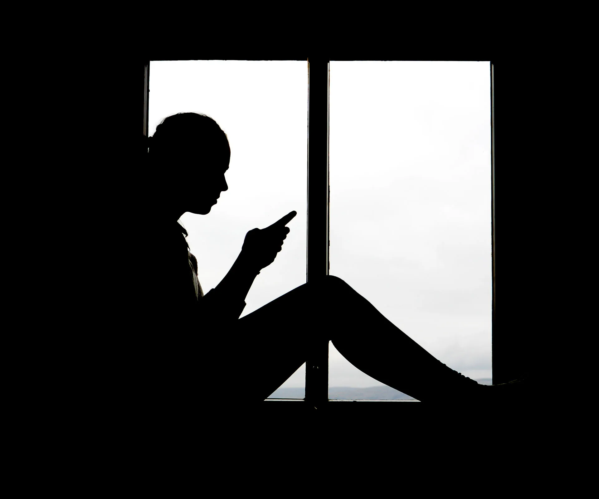 Silhouette of a person sitting by a window, looking at a smartphone.