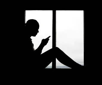 Silhouette of a person sitting by a window, looking at a smartphone.
