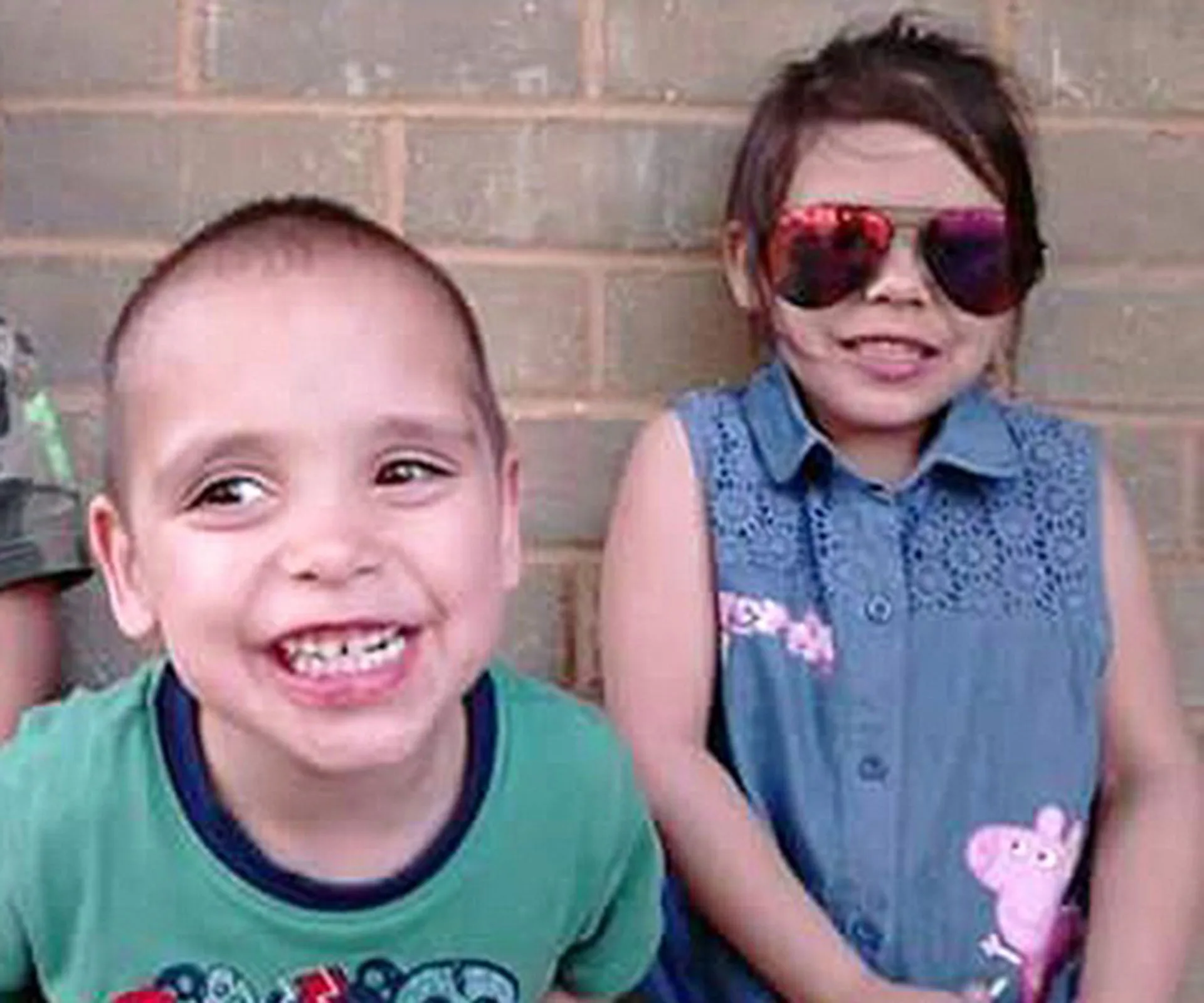 Two smiling children in front of a brick wall; boy in green tee, girl in denim dress wearing sunglasses.