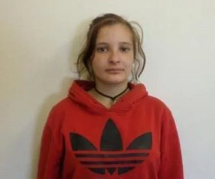 Young woman in red hoodie with black logo, straight hair, neutral expression, standing against plain background.