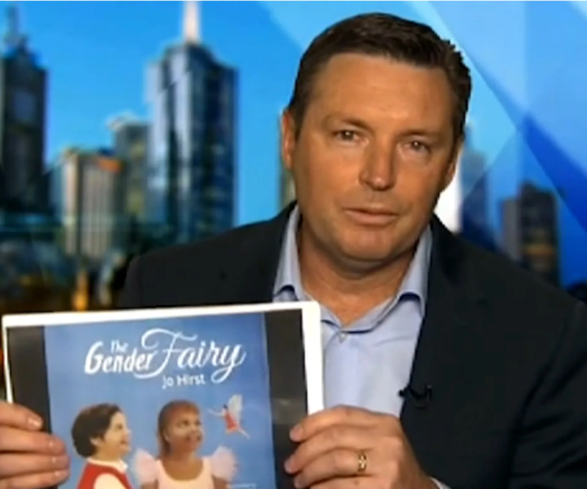 Man in suit holding "The Gender Fairy" book, with cityscape background.