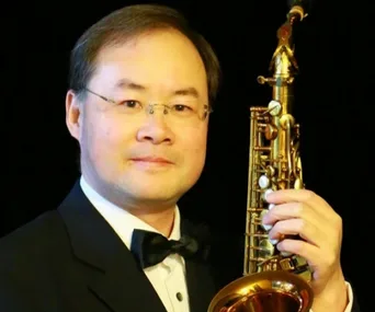 A man in a tuxedo holding a saxophone against a dark background.