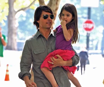 A man in sunglasses carries a young girl in a park. She wears a purple top and pink skirt, with trees in the background.