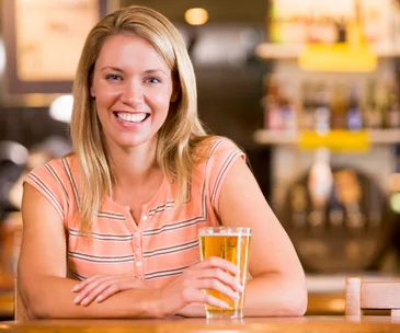 A woman smiling, seated at a bar, holding a glass of beer.