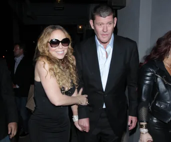 A woman in sunglasses and a black dress smiles, holding hands with a man in a suit, walking in a dimly lit setting.
