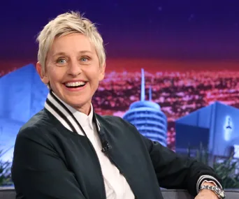 A person smiling broadly while seated on a talk show set with a cityscape backdrop.