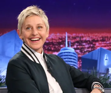 A person smiling broadly while seated on a talk show set with a cityscape backdrop.