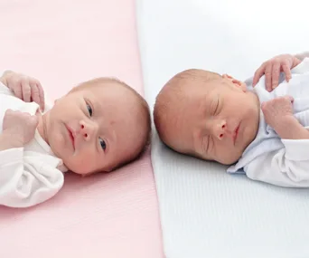 Two newborn twins lying on colored blankets, one awake and one asleep.