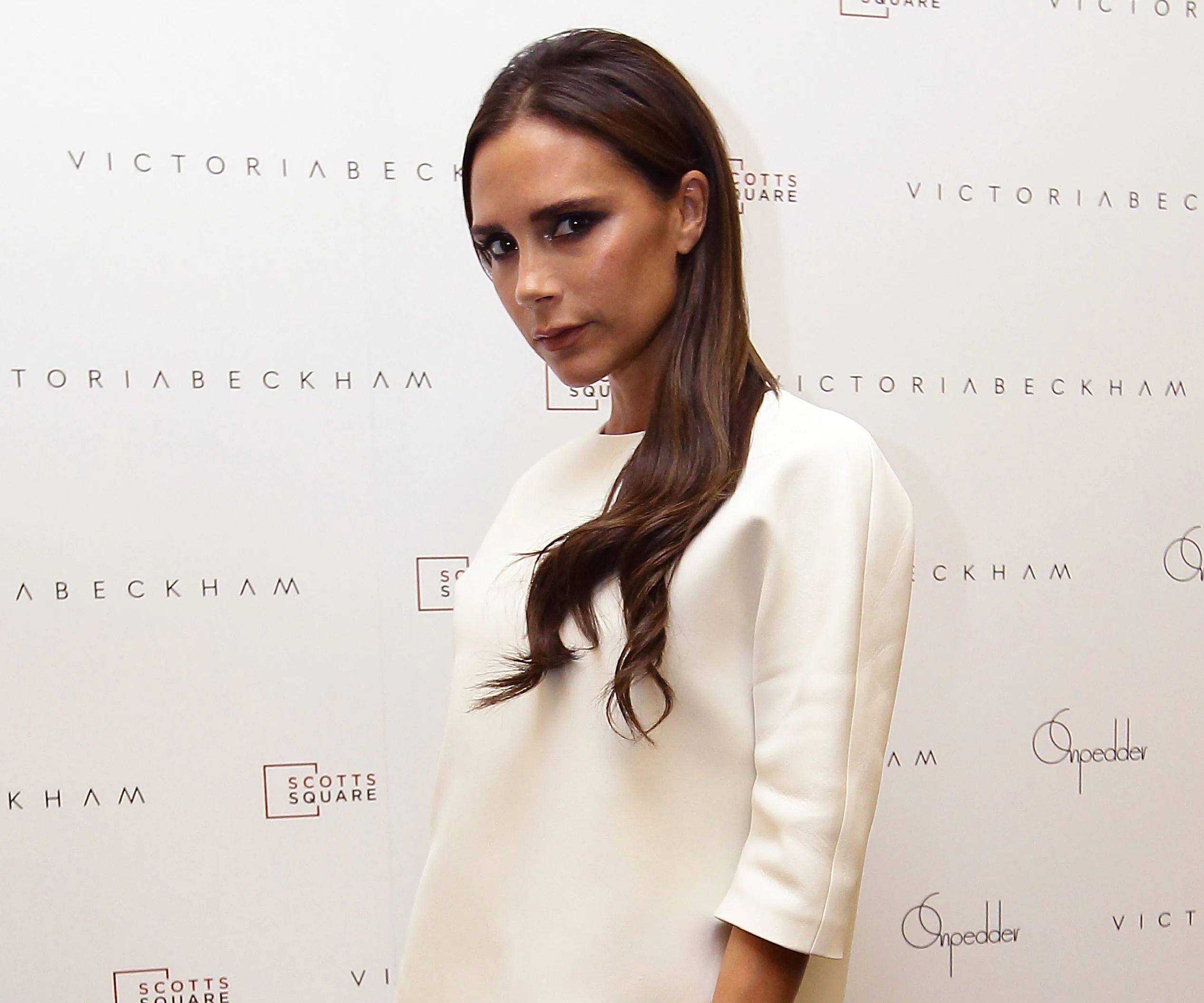 Victoria Beckham in front of a branded backdrop, wearing a white outfit, with long hair.