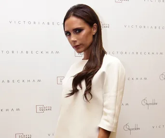 Victoria Beckham in front of a branded backdrop, wearing a white outfit, with long hair.