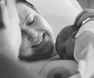 Mother smiling while breastfeeding her newborn baby, lying on a bed, depicted in a peaceful, intimate moment.