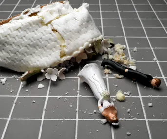 Broken wedding cake with toppled bride and groom figurines on gray tiled floor.
