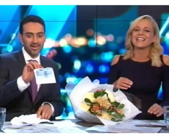 Presenter holds ultrasound photo, another smiles with flowers, on a talk show set.
