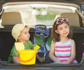 Kids sitting in the car trunk with beach toys and snorkeling gear, enjoying a sunny day trip.