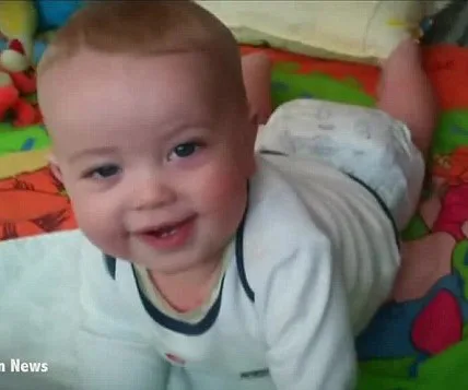 A smiling baby in a white outfit lies on a colorful play mat, looking towards the camera.