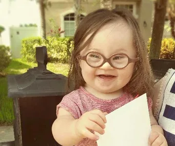 Child with Down syndrome wearing glasses, smiling and holding white paper while sitting outdoors on a bench.