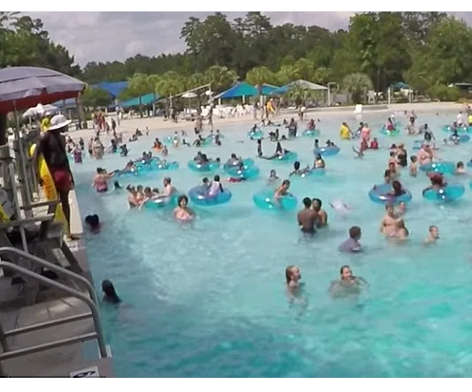 Lifeguard standing by a crowded pool, with people swimming and using inflatable rings on a sunny day.