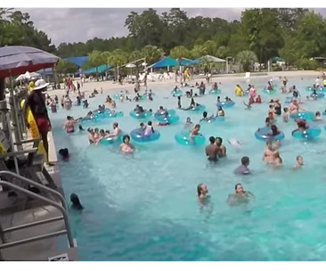 Lifeguard standing by a crowded pool, with people swimming and using inflatable rings on a sunny day.