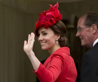 A woman in a red outfit and hat waves, smiling, with a man beside her.