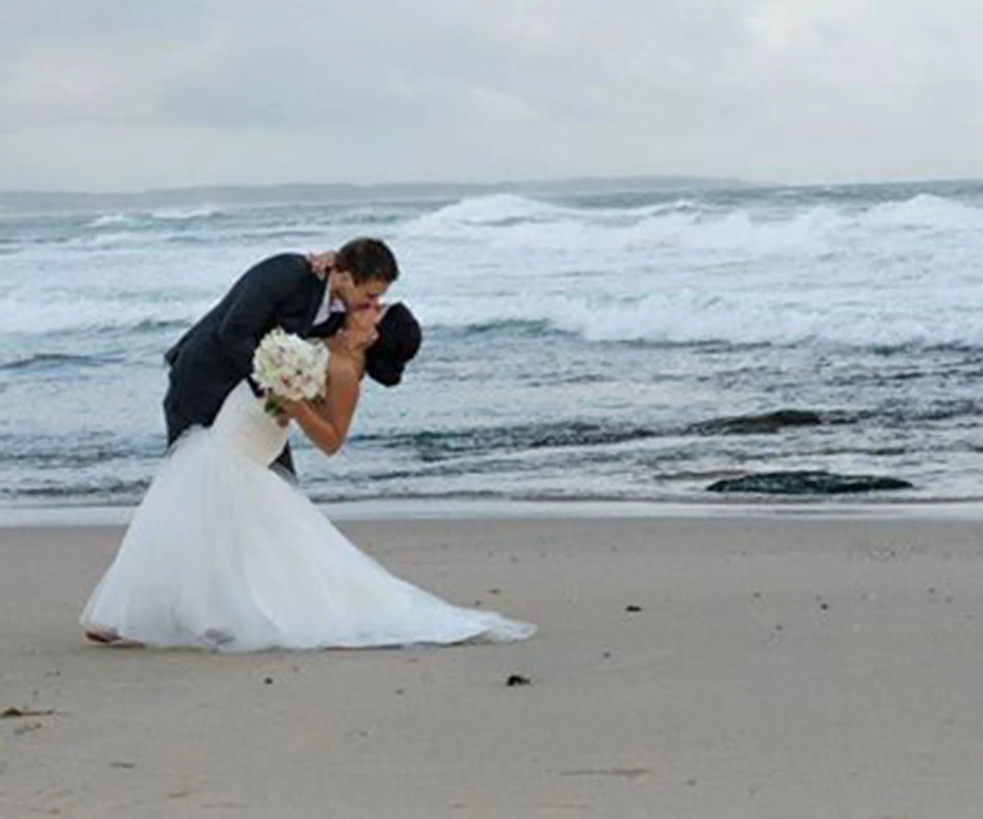 A newlywed couple shares a kiss on the beach, with waves gently crashing in the background.