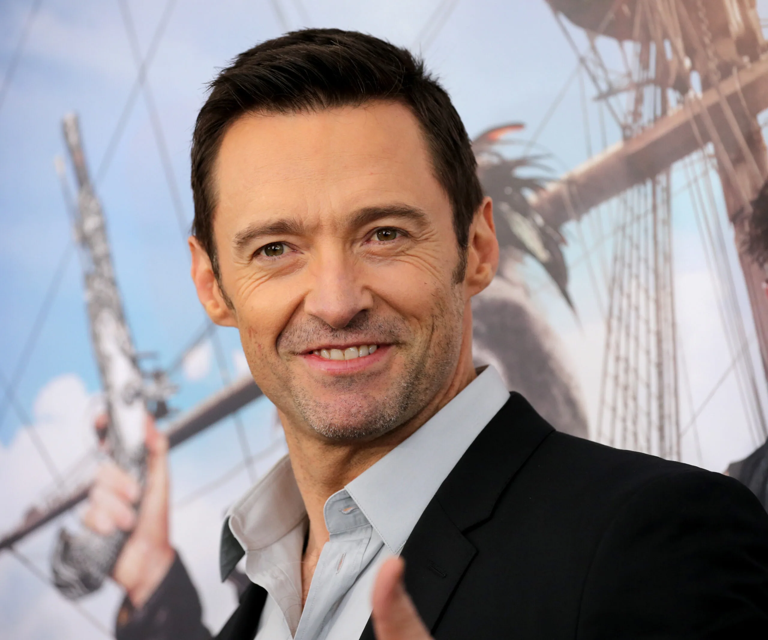 Hugh Jackman smiling at an event, wearing a dark suit with a light shirt, background shows part of a ship.