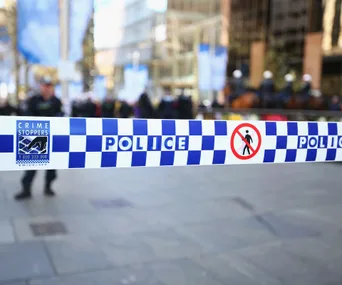Police tape with "CRIME STOPPERS" and "POLICE" at a city scene, blurred officers in background.