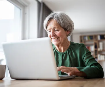 Senior woman in green shirt smiling while using a laptop at home.