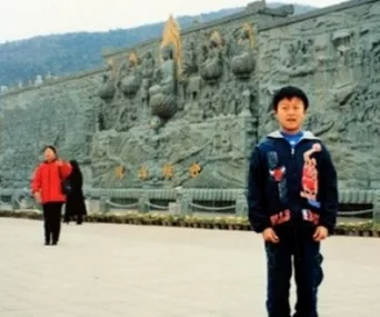 A young boy in a jacket poses in front of an intricate stone mural, with a woman in a red coat walking in the background.