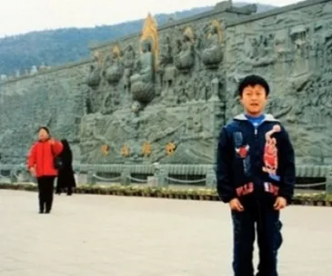 A young boy in a jacket poses in front of an intricate stone mural, with a woman in a red coat walking in the background.
