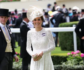 Woman in a white lace dress and hat, smiling at an outdoor event with people in formal attire in the background.