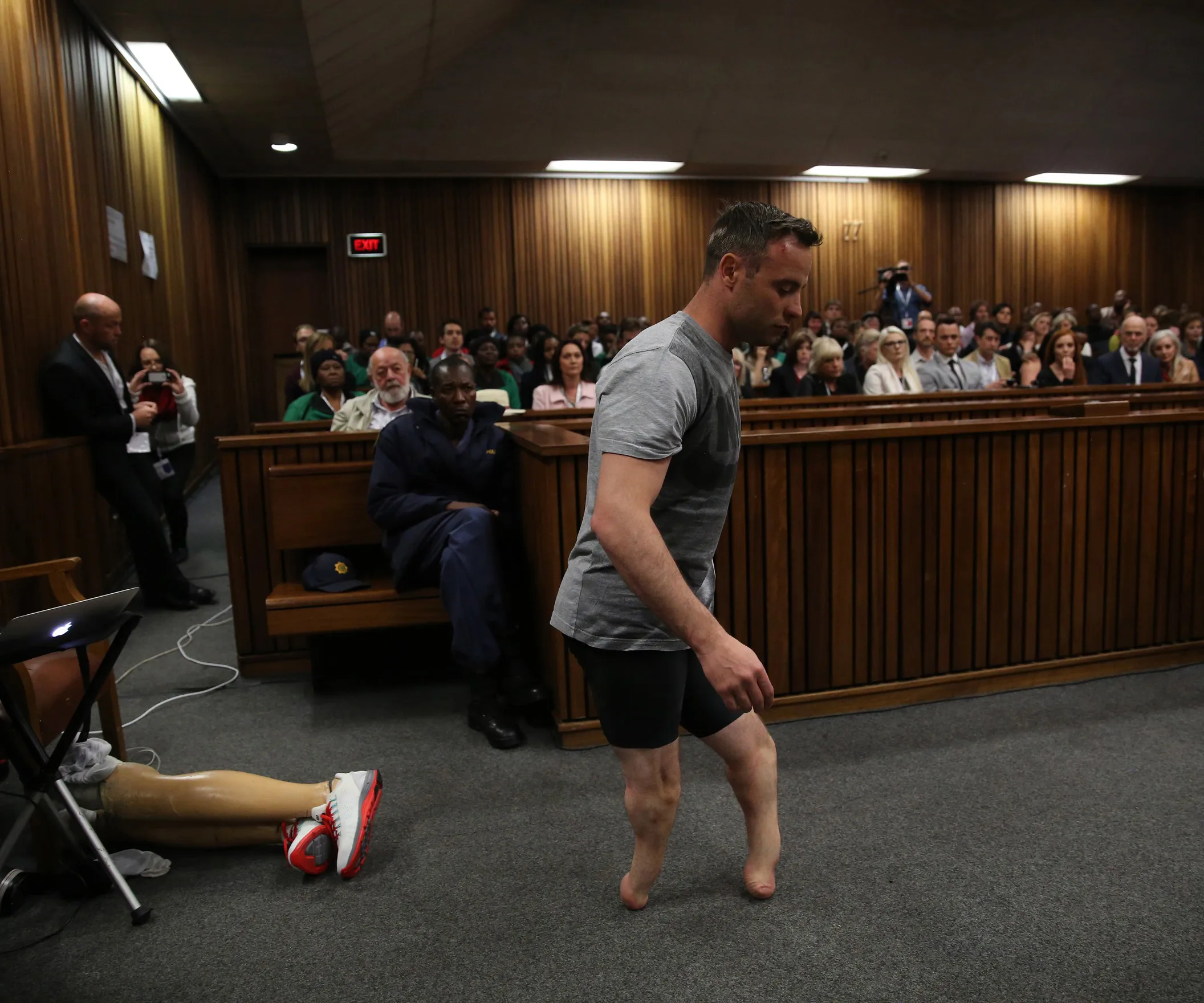 Man walks without prosthetics in courtroom to demonstrate vulnerability; audience and prosthetics visible in background.