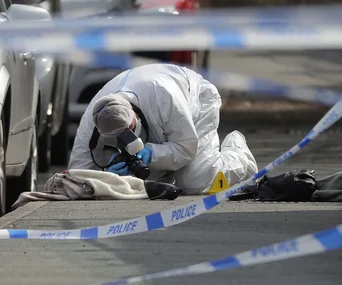 Forensic investigator examines a crime scene on a street, surrounded by police tape and evidence markers.
