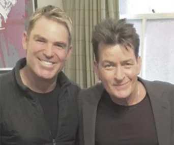 Shane Warne and Charlie Sheen smiling together indoors, standing close.