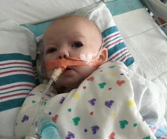 Baby in hospital bed with oxygen tube, wrapped in heart-patterned blanket.