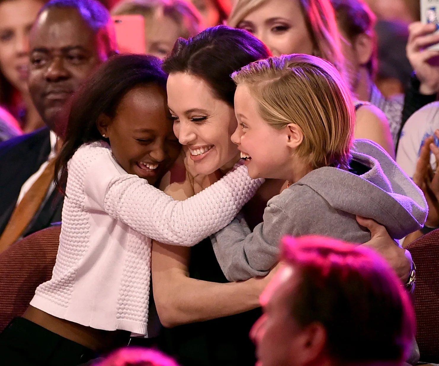 A woman joyfully hugging two children at an event, surrounded by an audience with cameras.