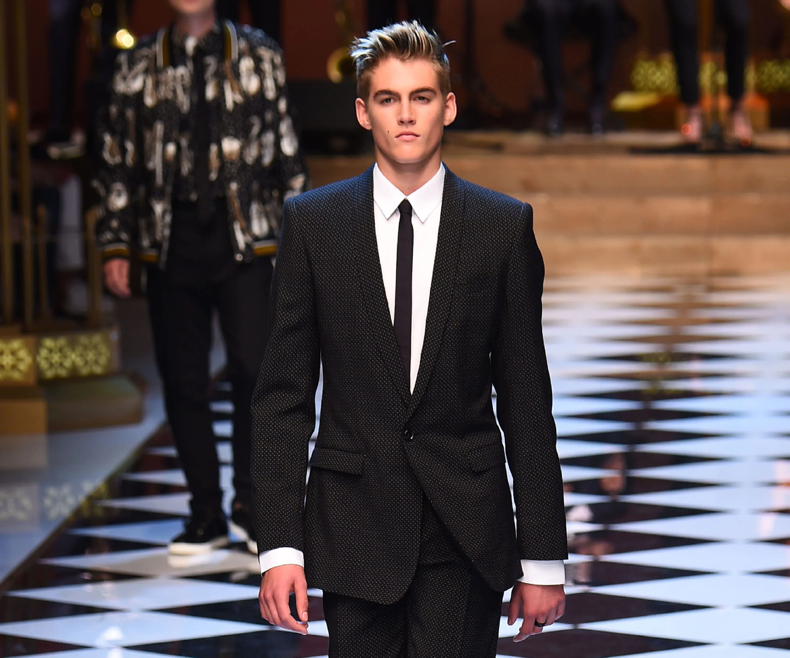 Model in a black suit and tie walks down a checkered runway confidently.