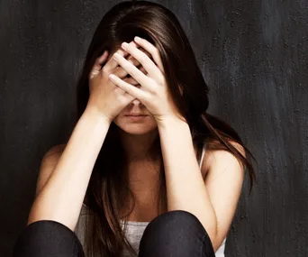 A woman sitting against a dark wall, covering her face with her hands, conveying distress or contemplation.