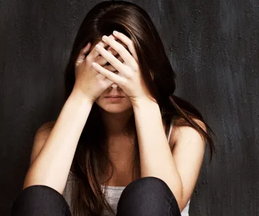 A woman sitting against a dark wall, covering her face with her hands, conveying distress or contemplation.