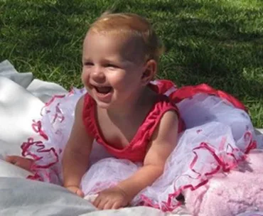 Toddler in a pink and white frilly dress smiles while sitting on a blanket in the grass.