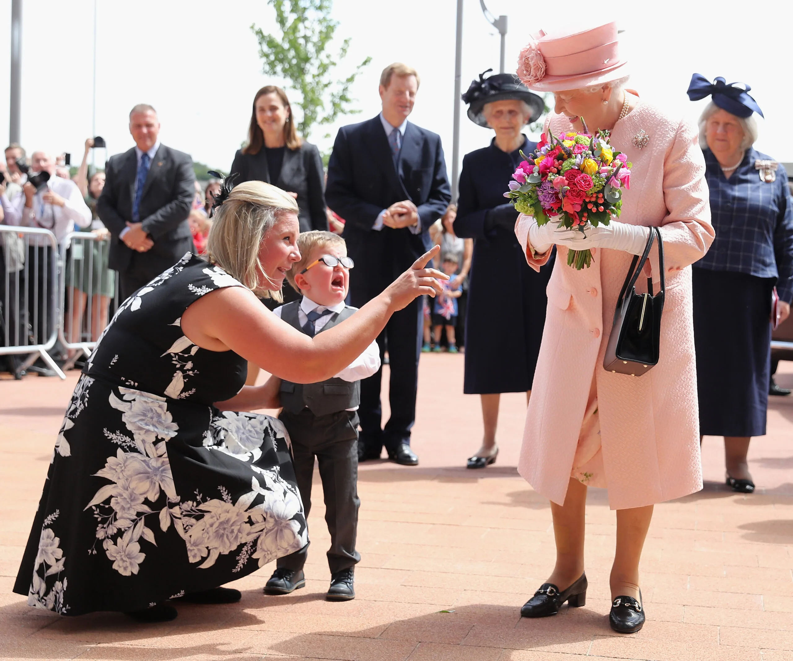 A young boy, visibly upset, meets an elderly woman in a pink outfit holding flowers. A woman is comforting the boy.